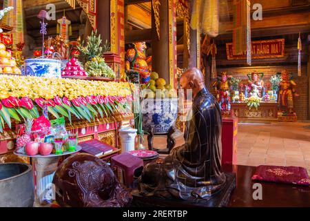 Polierte Bronzestatue eines Mönchs, der vor einem reich dekorierten Altar mit ernteten Lebensmitteln in einem buddhistischen Tempel auf dem Berg Fansipan, Sapa, kniet Stockfoto