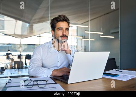 Ein aufmerksamer junger Geschäftsmann aus Lateinamerika, der wegblickt und denkt, er verwende bei der Arbeit ein Notebook. Stockfoto