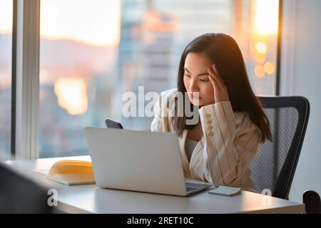 Junge gestresste Geschäftsfrau aus Asien, die im Büro Mobiltelefon benutzt. Stockfoto