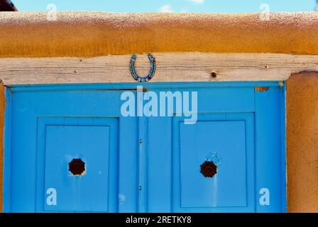 Ranchos de Taos, New Mexico, USA - 23. Juli 2023:Ein dekoriertes Hufeisen hängt über den blau bemalten Holzdoppeltüren eines traditionellen Hauses aus lehmziegeln. Stockfoto