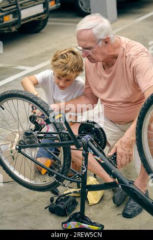 Der ganze Körper eines Großvaters, der Fahrradreifen aufpumpt, in der Nähe des Enkels, der mit Aufmerksamkeit auf das Rad schaut Stockfoto