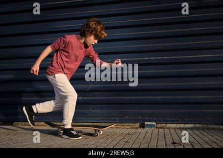 Ganzkörperjunge in legerer Kleidung läuft, während er auf dem Skateboard gegen die dunkelblaue Wand auf der Straße der Stadt reitet Stockfoto