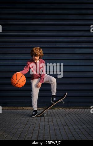 Ganzkörper-Junge auf dem Skateboard, der Basketball gegen die dunkelblaue Wand auf der Stadtstraße spielt Stockfoto