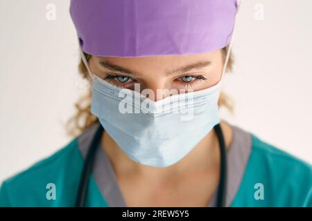 Besorgte Ärztin in medizinischer Uniform mit Stethoskop trägt Schutzmaske und blickt auf beigen Hintergrund hinab Stockfoto