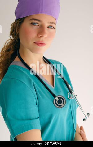 Selbstbewusste junge Frau in medizinischem Bademantel und mit Stethoskop faltbaren Händen und Blick auf die Kamera auf beigem Hintergrund Stockfoto