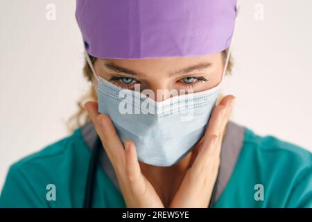 Professionelle Ärztin in medizinischen Peelings tragen Schutzmaske und berühren Wangen auf beigem Hintergrund Stockfoto