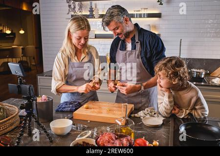 Niedliches Kind, das Eltern beim Schälen roher Kartoffeln am Tisch beobachtet und beim Kochen in einer modernen, leichten Küche einen Vlog aufnimmt Stockfoto