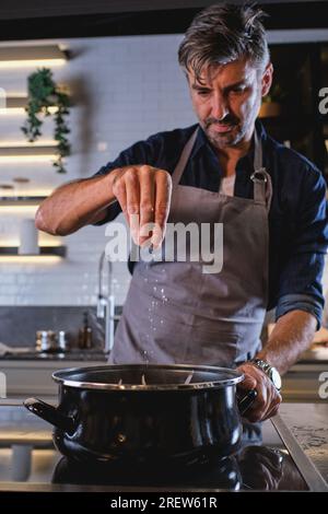 Bärtiger Mann in grauer Schürze, der Salz in den Topf schüttet, während er in der modernen Küche Essen zubereitet Stockfoto