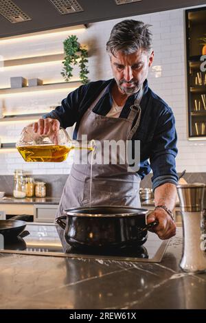 Fokussierter, bärtiger Koch in einer Schürze, der Öl aus der Flasche in den Topf gießt, während er in einer hellen, modernen Küche kocht Stockfoto
