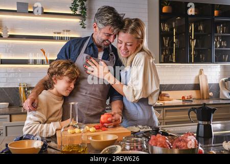Fröhlicher Mann mit Tomate in der Hand, umarmte Frau und Sohn, während er zusammen in der Küche Krake kochte Stockfoto