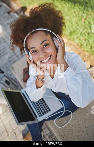 Der große Winkel einer Freiberuflerin mit Kopfhörern, die in die Kamera schaut und Musik hört, während sie im Stadtpark per Fernzugriff auf einem Netbook arbeitet Stockfoto