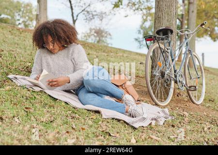 Konzentrierte ethnische Frau mit Brille, die auf einer Decke neben dem Fahrrad ruht und am Wochenende im Green Park interessante Romane liest Stockfoto