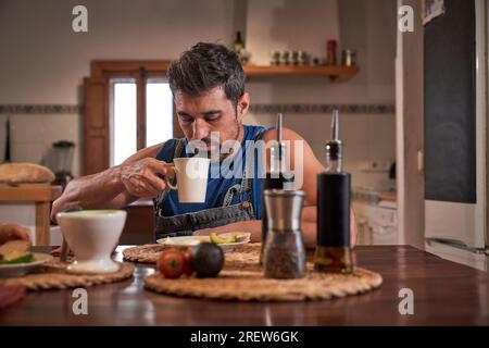 Seriöser bärtiger Mann, der am Holztisch sitzt und eine Tasse frischen heißen Kaffee serviert, während er morgens ein gesundes Frühstück hat Stockfoto