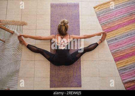 Luftaufnahme einer blonden Frau in Sportkleidung beim Yoga Während sie ihre Beine in einer Matratze auf dem Boden streckte Eines Hauses Stockfoto