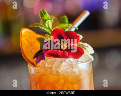 Abschluss eines erfrischenden Longdrink-Cocktails mit essbaren roten Blüten und getrockneten Orangenscheiben, serviert in der Bar auf verschwommenem Hintergrund Stockfoto