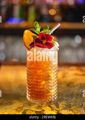 Longdrink-Cocktail mit Eiswürfeln, dekoriert mit Blumen und getrockneten Orangenscheiben, serviert in einer modernen Bar mit verschwommenem Hintergrund Stockfoto