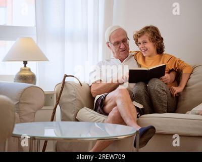 Ein sehr zufriedener älterer Mann mit positivem Enkel liest zusammen ein Interessantes Buch, während er auf dem Sofa in einem hellen Wohnzimmer zu Hause sitzt Stockfoto