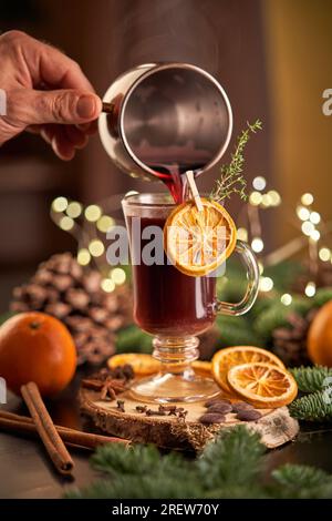 Ernte anonyme Person, die während der Weihnachtsfeiertage alkoholischen Glühwein mit Orangenscheibe auf dem Tisch mit Nadelzweigen in Glas gießt Stockfoto