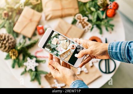 Draufsicht einer anonymen Person, die ein Foto von eingepackten Weihnachtsgeschenken auf dem Handy in der Nähe von Tannensprigs und Papier neben dem Wort xmax und Candy c macht Stockfoto