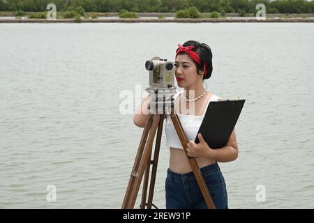 Hübsche asiatische Vermessungsfrau oder Ingenieurin, die mit Theodolith-Transitgeräten auf einer Baustelle im Freien arbeitet. Stockfoto