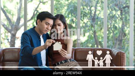 Ein junges asiatisches Paar sitzt auf einer gemütlichen Couch und steckt Silbermünzen in eine hölzerne, hausförmige Münzbox, die einen Hinweis auf Träume einer idyllischen Zukunft gibt. Stockfoto