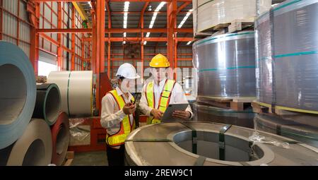 Industrielle Lagerhausszene mit zwei sicherheitsbewussten Männern, die Stahlblechrollen inspizieren, mit dem rauen Kern der täglichen manuellen Arbeit und Stockfoto