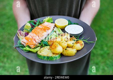 Frauenhände halten gebackenes Lachsfilet mit Kartoffeln und Gemüse in der Gartensaison. Stockfoto