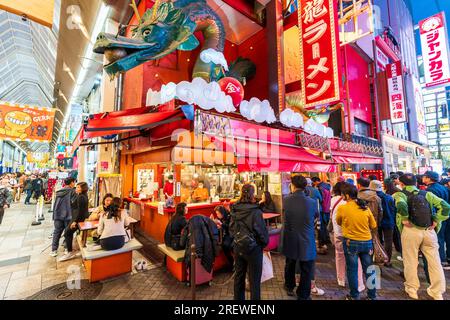 Das beliebte Restaurant Kinryu Ramen in Doronbori, Osaka, ist abends geöffnet. Eckgebäude mit Gästen, die draußen an Tischen essen, während andere sich um einen Platz in der Schlange anstellen. Stockfoto