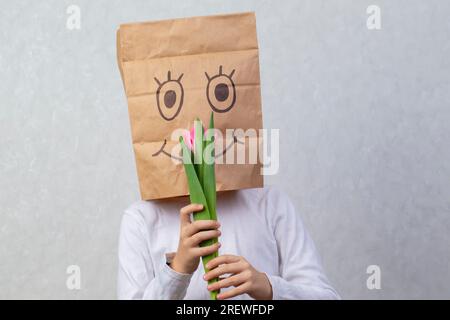 Ein Kind mit einer Pappschachtel auf dem Kopf hält eine Tulpe. Das Konzept eines anonymen Geschenks. Stockfoto