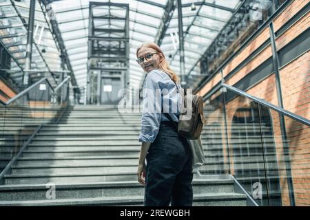 Glückliche Geschäftsfrau oder Studentin mit Rucksack, die die Treppe in einem modernen Saal im Loft-Stil erklommen hat. Konzept „Weg zum Erfolg“ Stockfoto