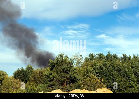 Schwarzer Rauch von brennenden Waldbäumen und Gebäuden vor einem blauen Himmel, ein Feuer mit viel schwarzem dunklem Rauch vor einem blauen Himmel mit Wolken Stockfoto