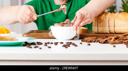Kind mit Schokoladenpaste und einem Laib, ein Kind bereitet in der Küche Süßigkeiten aus einem Brötchen und Schokolade zu, Schokoladenbutter, die das Kind auf eine Loa setzt Stockfoto