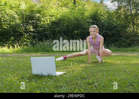 Ein Teenager-Mädchen ist im Park in Fitness verwickelt. Führt Übungen online mit einem Computer durch Stockfoto