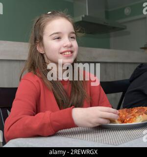 Ein Teenager isst Pizza. Porträt eines lächelnden Mädchens beim Abendessen in einem roten Pullover. Quadratisches Format Stockfoto