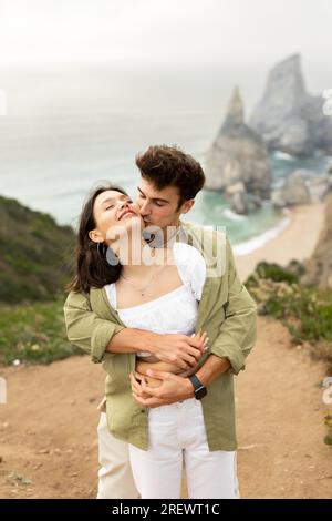 Ein hübsches junges europäisches Paar genießt ein Date an der Küste, ein Mann umarmt die Dame von hinten und küsst sie, vertikaler Schuss Stockfoto