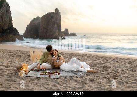 Romantische Ehepartner, die ein Picknick machen, auf einer Decke sitzen und sich umarmen, sich am Strand treffen und romantische Momente feiern Stockfoto