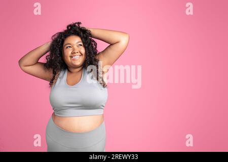 Portrait einer glücklichen, fetten afroamerikanischen Frau in Sportbekleidung, die über rosa Hintergrund posiert und auf den Kopierbereich schaut Stockfoto