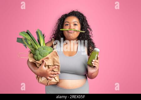 Witzige schwarze Dame in Sportkleidung mit Gemüsetasche und Flasche Smoothie, die Spaß im pinkfarbenen Studiohintergrund hat Stockfoto