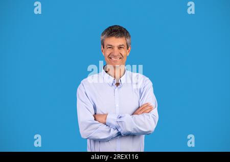 Porträt eines selbstbewussten, reifen Geschäftsmannes, der Hände über blauen Hintergrund kreuzt Stockfoto