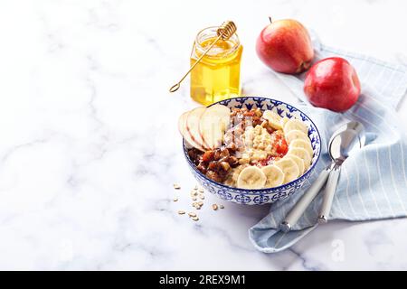 Haferbrei mit karamellisierten Äpfeln mit Zimt, Banane, geriebenen Erdbeeren und Honig auf hellem Marmorboden Stockfoto