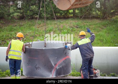 Arbeiter arbeiten auf der Baustelle. Ein Hydraulikbagger verwendet eine Kette, um ein Betonrohr anzuheben und einen Regenwasser- oder Kanalisationssammler zu installieren Stockfoto