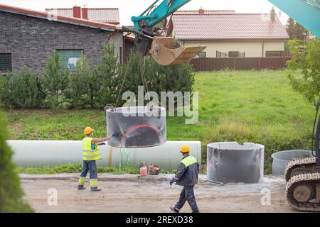 Arbeiter arbeiten auf der Baustelle. Ein Hydraulikbagger verwendet eine Kette, um ein Betonrohr anzuheben und einen Regenwasser- oder Kanalisationssammler zu installieren Stockfoto