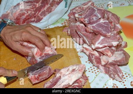 Hände eines Mannes, der ein großes Stück rohes Fleisch mit einem scharfen Messer in kleinere Stücke schneidet. Stockfoto