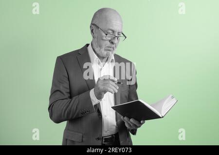 Porträt eines älteren Geschäftsmannes in einem Anzug mit Notizbuch und Stift denkt über die Aufgabe nach - auf grünem Hintergrund. Schwarz-Weiß-Konzept Stockfoto