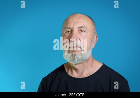 Porträt eines älteren Mannes mit grauem Bart in Freizeitkleidung. Schwarzes T-Shirt, blauer Hintergrund Stockfoto