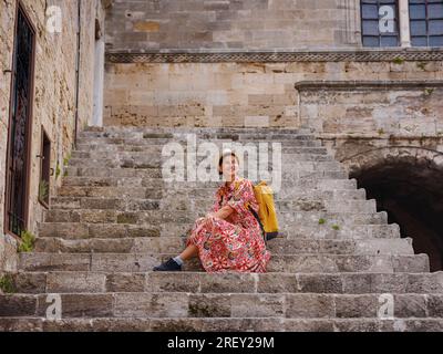 Junge Asiatin in rotem Kleid läuft durch die Straßen von Rhodos. Eine Frau, die sich auf alten Treppen in der Festung Rhodos ausruht. Tourismus, Urlaub und Entdeckungen Stockfoto