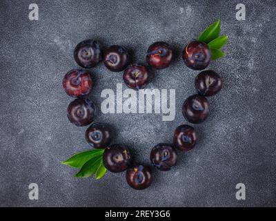 Süße Pflaumen auf dunklem Hintergrund. Frische blaue Pflaumen mit Blättern. Kulinarische Frucht im Hintergrund. Sommerpostkarte. Bei der Ernte im Garten liegen Pflaumen in Form von Gehör Stockfoto