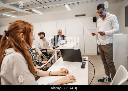 Generationenübergreifende Gruppe kreativer Menschen, die eine Präsentation im Klassenzimmer halten. Männlicher Manager mittleren Alters, der Monatsfinanzbericht liest Stockfoto