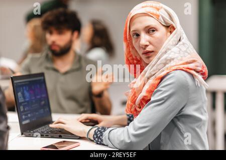 Schöne Frau in Hijab, abgelenkt von etwas, während sie auf dem Laptop arbeitet und etwas ansieht Stockfoto