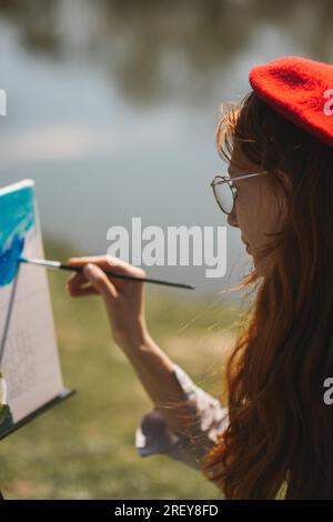 Nahaufnahme eines Ingwer-Mädchens mit Brille und roter Baskenmütze auf einer Leinwand auf einer Staffelei, während Sie im Park in der Nähe des Sees sitzen Stockfoto